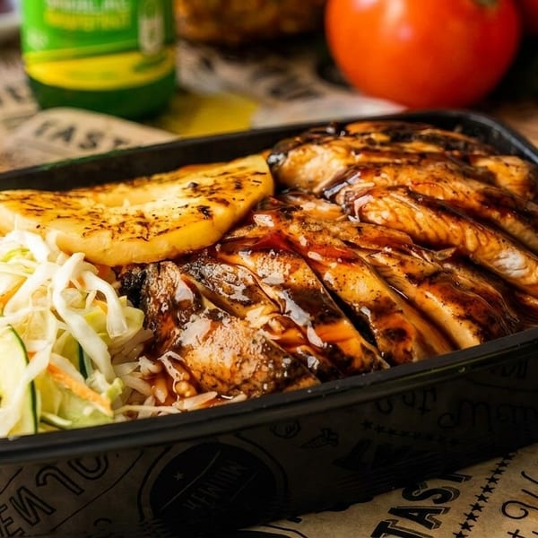 Grilled chicken with sauce, pineapple, and coleslaw in a black to-go container with a soda.
