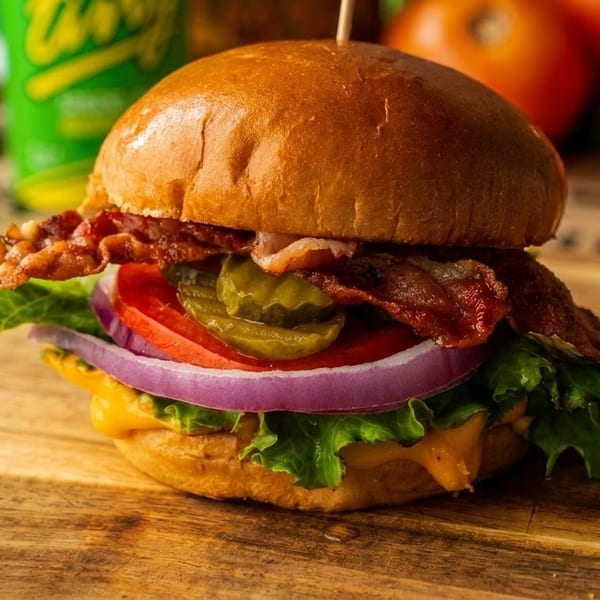 Close-up of a bacon cheeseburger with lettuce, tomato, onion, and pickles on a wooden board.