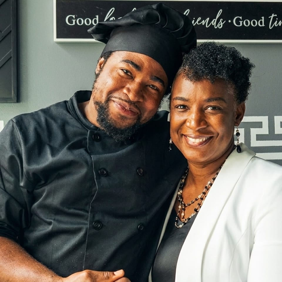 A smiling chef in a black uniform and a woman in a white blazer pose together.