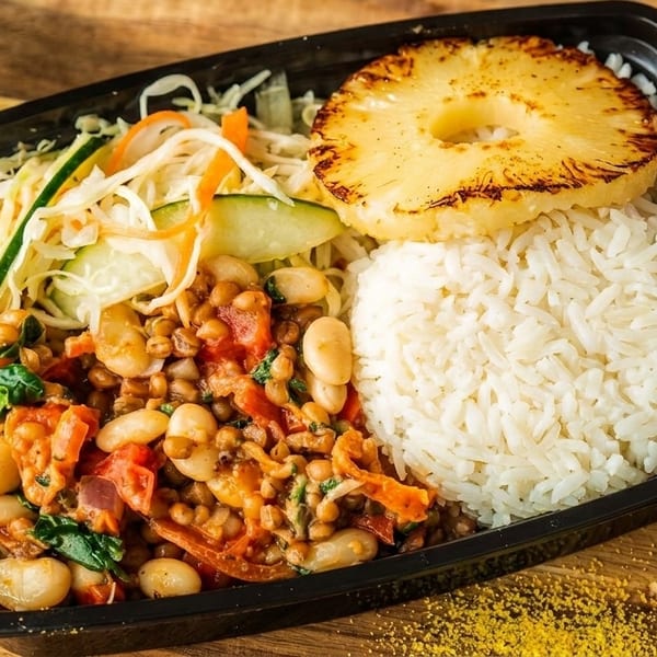 A black takeout container filled with rice, grilled pineapple, bean stew, and shredded salad.